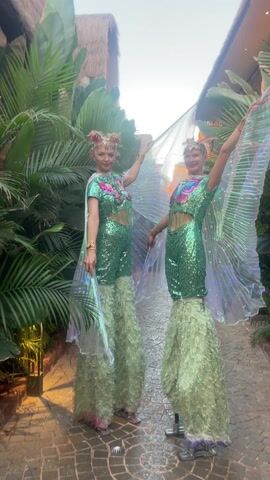 Two women in elaborate green, sequined costumes and stilt extensions pose outdoors on a brick-paved path lined with tropical foliage. They are wearing matching outfits with short-sleeved tops, midriff cutouts, and sheer capes that resemble wings. Their faces are painted, and they wear ornate headpieces. Both women have their arms raised and are smiling. In the background, there are thatched-roof buildings and glimpses of other individuals. The setting appears to be a tropical or exotic location, possibly a resort or event space.