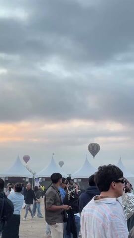 A crowd of people is gathered outdoors. Several white tents are visible in the background. Hot air balloons are floating in the sky. Most of the people are facing towards the balloons, and some are taking photos or videos with their phones. A man in sunglasses and a striped shirt is in the foreground. The sky is cloudy.