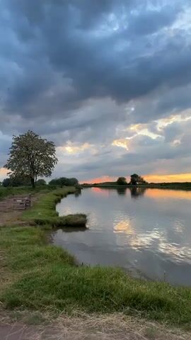The video captures a serene sunset over a lake or large pond in Doganovo, Bulgaria. The sky is overcast with dramatic clouds, yet vibrant orange and yellow hues from the setting sun pierce through, reflecting beautifully on the calm water.

The scene features lush green grass and reeds in the foreground and along the banks. Trees line the far side of the water body and are also visible on the left bank closer to the viewer.

The video is filmed from inside a moving vehicle. One shot shows the wide expanse of the lake and sky, another focuses through the car's side window with the side mirror reflecting the dirt road, and a third shot provides a view through the windshield, showing the unpaved road ahead, flanked by tall vegetation and trees, leading towards the distant sunset. The time of day is clearly evening, characterized by the golden light of the setting sun. No people or visible text are present.