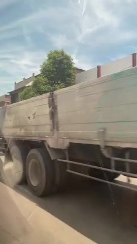 The video shows a view from a vehicle traveling on a road with buildings and trees on the side. Several vehicles are visible, including a large truck with a silver cargo box, a dark minivan, a yellow and red tanker truck, and a white SUV. The sky is partly cloudy with a bright sun. The road appears to be paved. The buildings on the side of the road seem to be commercial establishments, with some under construction. The video captures movement and activity, suggesting a daytime scene.