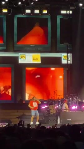 The video shows a male performer on a stage, wearing a red t-shirt and blue shorts. He is in the center of the stage, singing and dancing. There is a smoke machine emitting white smoke from the right side of the stage. In the background, there are large screens displaying abstract colorful graphics, primarily red, blue, and yellow. The screens are framed to resemble old television sets. There are also what appear to be Best Buy logos and a price tag displayed on the screens. The performer is lit by stage lights and there is an audience visible in the foreground, out of focus. Another person, possibly a musician, can be seen on the right side of the stage behind the performer, near a drum set. The overall atmosphere is that of a live music concert or performance.