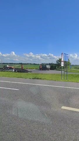 A car drives on an asphalt road with white markings, passing a blue road sign with white text, a street light, and a roundabout sign. The car continues along the road, with a large green field and a blue sky with white clouds visible in the background.
