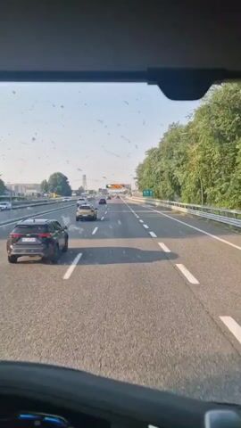The video is taken from inside a vehicle, looking forward through the windshield. The vehicle is traveling on a multi-lane highway. There are several other cars on the highway, all traveling in the same direction. The highway is bordered by trees and vegetation on the right side. There is a clear sky, suggesting daytime. A digital highway sign is visible in the distance. The windshield has several specks of dirt on it.