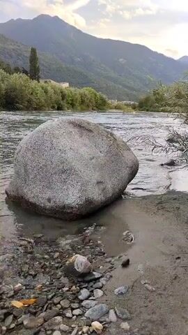 A wide shot shows a river flowing past a large gray boulder on the bank. Trees and hills are visible in the background, with a cloudy sky overhead. The water is clear and the riverbed is rocky. The boulder is smooth and worn, suggesting it has been in the river for a long time. The scene is peaceful and serene, with a sense of natural beauty.