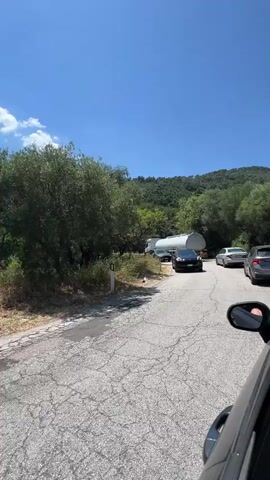 This is a first-person perspective video taken from inside a vehicle. The camera is looking out the side window, showing a winding asphalt road. Several cars are visible on the road, including a silver SUV, a dark-colored sedan, and a light-colored sedan further ahead. In the distance, a large tanker truck is navigating a curve on the road. The surrounding environment is rural and hilly, with lush green trees and vegetation on either side of the road. The sky is bright blue with a few white clouds, indicating a sunny day. The road appears to be in moderate condition, with some cracks visible in the asphalt. The overall scene suggests a scenic drive through a natural landscape, possibly in a mountainous or hilly region.