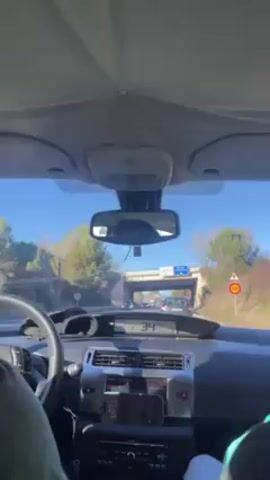 The video shows a car driving on a sunny day toward a small bridge. The speed is decreasing, from 34 km/h to 30 km/h. The dashboard is visible, including a smartphone mounted on a holder. Trees line the side of the road. The view is from the driver's perspective.