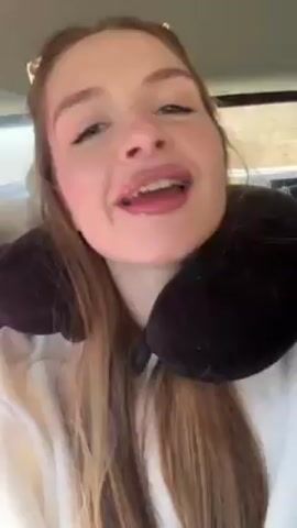 The video shows a young woman sitting in a vehicle, wearing a black travel pillow and a white sweatshirt. She is holding up her hand, displaying bright red nail polish. She is smiling and singing along to music that is playing in the background. The interior of the car is visible, with a pink object and some other items behind her. The scene appears to be casually recorded, possibly as a short video for social media.