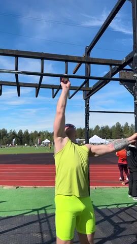 The video shows a man wearing a bright yellow tank top and shorts standing beneath a black metal outdoor workout structure. He is fair-skinned and has a beard and tattoos on his arms. He reaches up with one hand to grip a bar on the structure, looking upward. There are people in the background, along with a red running track, a green grassy area, trees, and a blue sky with some clouds.