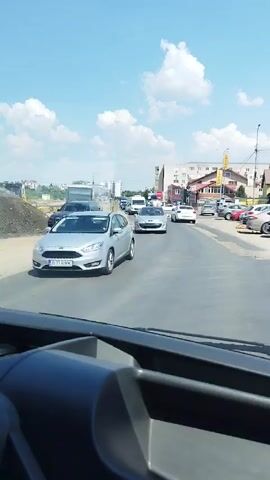 A video captures a view from inside a vehicle, looking out at a road scene on a sunny day. The road appears to be undergoing construction or repair, with visible patches of uneven asphalt and dirt on the sides. Several cars and a white van are driving on the road. On the right side of the road, there are buildings with shops and signs. The sky is blue with scattered white clouds. The perspective suggests the video is taken from the driver's seat or a passenger seat of a vehicle.