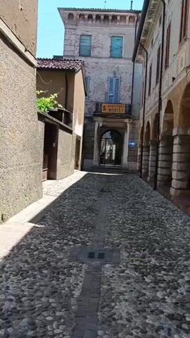 The video shows a long, narrow alleyway paved with cobblestones. The alleyway is flanked by old buildings. On the right side, a building with arched walkways supported by columns lines the street. On the left, the building has a simple, unadorned wall. At the end of the alleyway, another building stands with a sign reading "MANTOVANI MOBILI" above its entrance. A blue directional sign with a white arrow pointing left is fixed to the wall near the entrance. Sunlight casts shadows across the cobblestone street. The weather appears to be sunny. No people are visible in the video.
