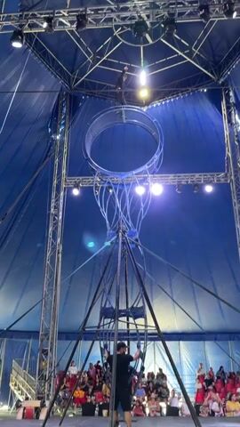 A performer stands on top of a large metal wheel structure inside a blue circus tent. The performer is wearing a black outfit and a black hat. The wheel is suspended high above the ground by a metal frame. The performer is looking to the left and appears to be preparing for a performance. Lights are visible on the tent ceiling and around the wheel structure. The overall environment suggests a circus performance is about to begin or is in progress.