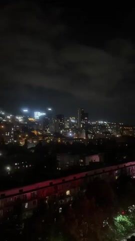 This is a vertical video shot at night from a high vantage point overlooking a city. The video pans from left to right, showing various buildings illuminated by lights. In the foreground, there are trees with some green foliage. Further in the distance, the city is spread out with numerous buildings, some tall with illuminated windows and facades, others lower-rise apartment buildings. There are also streetlights visible, and the overall impression is of a lively urban landscape at night. The sky is dark with some clouds. The camera movement is smooth, creating a sweeping view of the cityscape.