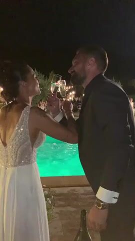 A man and a woman are standing next to a pool at night. The woman is wearing a white dress with sequins on the bodice and the man is wearing a black suit. They are both holding champagne glasses and appear to be toasting each other. In the background, there are lights strung up and other people can be seen. A bottle of champagne is on the ground in front of them.