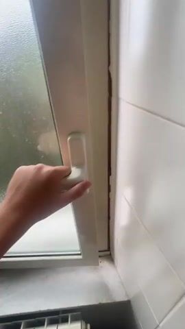 A hand is holding open a white window frame revealing a view of trees and mountains outside. The interior is tiled with white tiles and a windowsill with what appears to be marble is visible. The sky outside is cloudy.