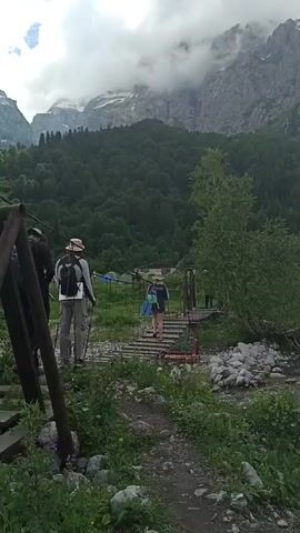 The video shows a group of people crossing a suspension bridge over a rocky stream. The bridge is made of wood and has metal supports. The people are wearing backpacks and appear to be hiking. The environment is mountainous, with lush green trees covering the slopes. The sky is cloudy, and the mountains in the background are partially obscured by mist. There are tents set up in the background, suggesting a campsite. The camera is positioned on the side of the path leading to the bridge, focusing on the people as they cross. The lighting suggests it is daytime, likely overcast or early morning/late afternoon.