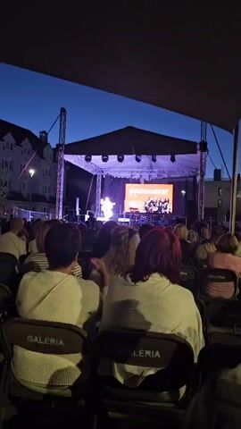 The video shows an outdoor concert at dusk. A man is playing an acoustic guitar on a stage, wearing a colorful shirt and shorts. A large screen behind him displays text that appears to be Hungarian, along with a group photo. The stage is lit with purple and white lights, and there are speakers on either side of the stage. A crowd of people are seated in chairs facing the stage, with their backs to the camera. The chairs have the word "GALERIA" printed on them. The sky is dark blue, indicating evening.