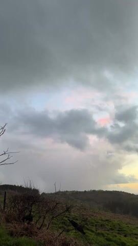 The video pans across a landscape under a dramatic sky. The foreground features sparse shrubbery and patches of green grass. The midground consists of rolling hills covered in trees, leading to a valley with visible buildings and possibly a small body of water. The sky is filled with grey clouds, but there is a strip of orange and yellow along the horizon, suggesting a sunset or sunrise. The overall atmosphere is tranquil but slightly somber due to the heavy cloud cover.