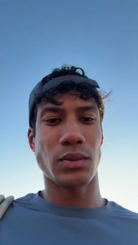 The video begins with a close-up shot of a man's face looking upwards. He is wearing a dark baseball cap and a grey t-shirt. The sky is visible in the background. The shot then shifts to a closer view of the man's face, showing him chewing. The camera then pans to the right, revealing a man standing on artificial grass in front of a wall with logos. In the background, there are people standing near a white structure shaped like a soccer ball. The video concludes with another close-up of the man's face, looking directly at the camera. The lighting suggests it is daytime, possibly late afternoon or early evening.