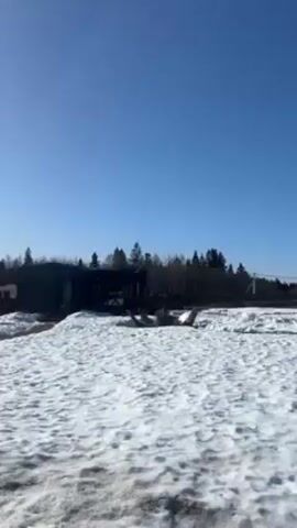 The video shows a snowy field on a bright, sunny day. In the background are dark buildings and a treeline. The snow is uneven and covers the ground completely. There are a few wooden planks or structures partially visible in the snow. The sky is clear blue. The camera seems to be static.