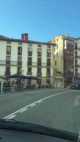 The video shows a street scene in a town. The perspective is from inside a vehicle, looking out through the windshield. On the left side of the street are several multi-story buildings with balconies and windows. There are awnings and tables set up outside one of the buildings, suggesting a cafe or restaurant. A few people are visible sitting at the tables. On the right side of the street, there are more buildings of similar architecture, with some trees and greenery visible further up the hill. Several vehicles are present on the street, including a dark-colored van, a silver van, and a dark car. The street has lane markings and a traffic light. The sky is clear and blue. The video captures a sense of movement as the vehicle drives along the curved street.