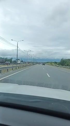 The video captures a view from inside a vehicle traveling on a highway. The highway has multiple lanes and is bordered by a guardrail on the left. Streetlights are visible along the side of the road, and power lines run overhead. Several cars and trucks are also seen on the highway in the distance, suggesting moderate traffic. The sky is cloudy. The interior of the vehicle, specifically the dashboard area, is visible in the foreground.