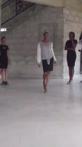 A medium shot captures three women in a spacious room. The central figure walks forward, wearing a silver top with shiny sleeves tied at the waist, a dark skirt, and heeled sandals. She pauses to pose with her hands on her hips. To her left, a woman stands wearing a black dress and black shoes. To her right, another woman in a similar black dress and white shoes stands with arms folded. The room has a light-colored, polished floor and a stone-textured wall. A staircase is partially visible in the upper left corner of the frame. The lighting appears natural.