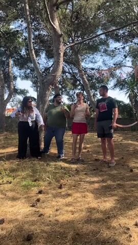 The video shows four people standing together in a grassy, wooded area. The first person on the left is a woman wearing a light-colored top, dark pants, and sunglasses. The second person is a man wearing a green shirt and jeans. The third person is a man wearing a white tank top and red shorts. The fourth person is a man wearing a dark t-shirt and grey shorts, holding a curved wooden object. They appear to be speaking or performing, although the audio is not analyzed. The ground is covered with pine straw and pinecones, and there are trees overhead with sunlight filtering through the branches. A hammock is visible in the background to the right.