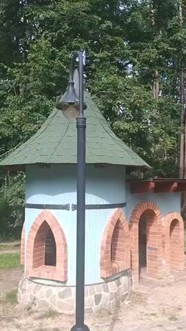 The video shows an outdoor scene with several small, castle-like structures made of wood, brick, and stone. The structures have conical roofs and arched entrances. There are trees and foliage in the background. Two young males are visible. One male, wearing a black t-shirt and green shorts, walks away from the camera and towards the castle structures. Another male, wearing a white t-shirt, sunglasses, and blue shorts, stands near the castle structures and appears to be gesturing. The ground is covered in dirt and paved stones. The lighting suggests it is daytime.