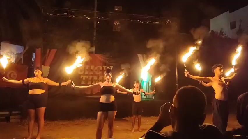 This is a video of an outdoor fire performance at night. In the foreground, a person is partially visible from behind, holding a phone up to record. In the background, there are five performers on a stage. From left to right, there is a woman holding a flaming torch in her left hand and her right arm extended. Next to her is another woman, also holding flaming torches in both hands, which are extended outwards. Behind them, a young boy is standing. To the right of the boy is a man with his arms extended, holding flaming torches. Finally, on the far right, another person is visible, holding a flaming torch. The stage has a banner behind it that says "SHOW TIME". There are lights and smoke effects visible. The ground appears to be sandy.