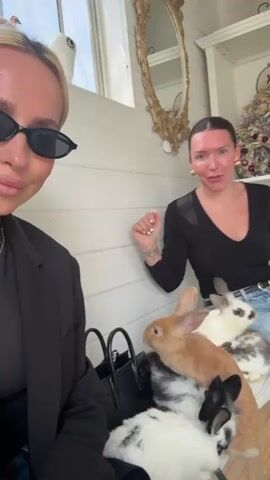 Two women are sitting on a bench inside a white, enclosed space, likely a shed or small building, surrounded by several rabbits. The woman on the left is wearing black sunglasses, a black blazer, and a gold chain. She is holding a black handbag and smiling at the camera. The woman on the right is wearing a black long-sleeved shirt and blue jeans. She has a surprised or excited expression and is holding her hand up. Several rabbits of different colors (white, brown, black and white) are gathered around them on the bench. The interior is decorated with a gold-framed mirror, a wreath, and shelves with decorative items. The background wall is white with horizontal planks. There's a window visible on the left side of the frame. Both women seem delighted by the presence of the rabbits.