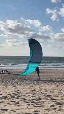 The video shows a beach scene. The foreground consists of a sandy beach with sparse vegetation. In the middle ground, a person stands behind a large turquoise and grey kite on the beach, which is partially lying on the ground. The kite is labeled "AIRUSH". In the background is the ocean with small waves breaking on the shore. The sky is blue with scattered white clouds. The overall lighting suggests it's daytime under a sunny sky. Another kite is visible in the far-left side of the screen.