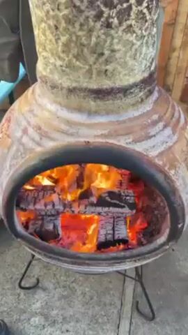 The video shows a lit chiminea outdoors. The chiminea is large and made of a weathered material, possibly clay or ceramic, with brown, cream, and gray patterns. It stands on a simple black metal stand with four legs. Inside the chiminea, there is a fire burning with bright orange flames and glowing red embers, and stacked wooden logs. A person wearing a gray jacket is visible on the left side of the frame, partially obscuring the view of the chiminea. Another person's legs and white shoes are visible near the base of the chiminea. In the background, there's a wooden fence or wall, and the ground appears to be concrete or paved.