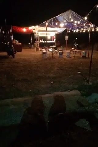 The video shows an outdoor food stall at night. A large tent is set up with string lights illuminating the area. Tables and chairs are arranged for customers. There is a counter where food is prepared. A motorcycle is parked to the left of the stall. The ground is covered in dirt and grass. The overall atmosphere appears to be casual and inviting.