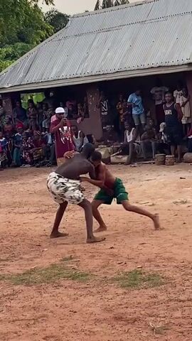Two men are engaged in a traditional wrestling match in an outdoor dirt arena in Achara, Nigeria. Both wrestlers are shirtless, with one wearing white and black patterned shorts and the other wearing green shorts. They are bent forward, heads pressed together, grappling for an advantage.

A large crowd of people of various ages surrounds the wrestling area, watching intently. Many are standing, while others are seated on wooden logs or benches near a building with a corrugated metal roof. One man in a maroon shirt and white cap stands near the wrestlers, appearing to oversee the match.

The setting suggests a community gathering in a rural or semi-rural area. The sky is overcast or it's late afternoon, providing soft, diffused lighting. Green trees, including palm trees, are visible in the background. The atmosphere is one of focused observation and engagement from the spectators.