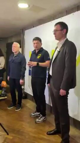 Three men stand side by side in an indoor setting, likely at a press conference. The man in the middle, dressed in a navy blue polo shirt with a yellow design, speaks with hand gestures. He stands on a wooden floor with a microphone clipped to his shirt. To his left, a man with gray hair wears a dark blue jacket and black pants. To the right, a man in a brown suit and glasses stands with hands at his sides. Behind the three men, there is a white banner with blue and yellow designs, suggesting a possible affiliation or organizational backdrop. The room appears to be simply decorated.