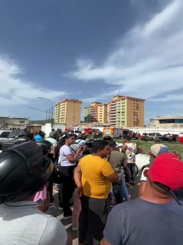 This media file captures a scene of destruction and emergency response in Turmero, Venezuela. The primary subjects are a large crowd of people gathered, observing the aftermath of what appears to be a building collapse. In the background, several tall apartment buildings stand, with one in the center showing significant structural damage, its lower floors completely demolished and a large pile of rubble accumulating at its base.

Emergency vehicles are present, including an ambulance with "EMERGENCIAS" and "AMBULANCIA DE EMERGENCIA" written on its side, and a fire truck with "ARAGUA" visible. The presence of these vehicles and the crowd suggests an ongoing emergency situation, likely a rescue or recovery operation. The people in the foreground are looking towards the damaged building, some appearing to be filming with their phones, while others are in conversation. The mood seems to be one of concern and observation.

The setting is outdoors, under a bright, clear sky with scattered clouds, indicating it is daytime. The presence of apartment buildings and what looks like a street or parking lot area places the scene in a residential or urban environment. The collapsed structure is the most prominent object, signifying a major event.

Notable details include the multi-colored facades of the undamaged apartment buildings, contrasting sharply with the devastation below. The rubble consists of concrete, rebar, and other building materials, indicating a catastrophic failure. The text visible on the emergency vehicles provides geographical context, identifying the emergency services as belonging to Aragua. The overall impression is one of a community responding to a disaster.
