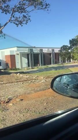 The video is a low-quality shot of a building taken from a moving car. The building appears to be a single-story conference center with a gray and white exterior. It features several large, dark-framed windows and vertical columns. The surrounding environment includes patchy green grass, red dirt, construction materials (bricks, sand), and a few trees. The sky is blue and clear. The car's side mirror is visible in the lower right corner of the frame. The video has some audio of people talking.