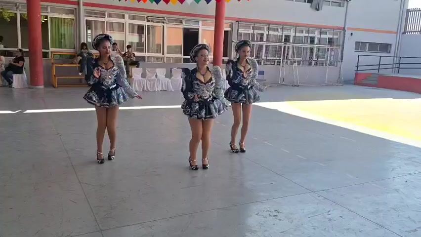 Three young women in matching sequined outfits and headdresses are dancing in a courtyard. They are positioned in a line, facing the camera, and appear to be performing. The courtyard has a concrete floor and is bordered by a building with large windows. In the background, there are people sitting and watching, as well as some playground equipment. A person holding a camera is visible in the lower right corner of the frame. The women are wearing high heels and their skirts are short and flared. The outfits are elaborate, with intricate patterns and shimmering details. They are all smiling and appear to be enjoying themselves. The overall atmosphere is festive and celebratory.