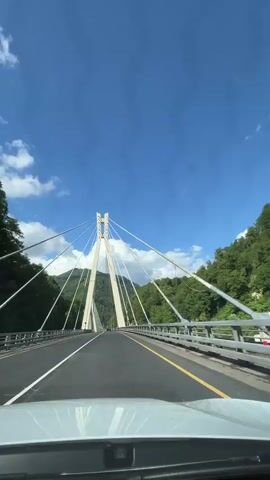 The video begins with a view from the driver's seat of a vehicle on a bridge. The bridge is a cable-stayed bridge with two distinct towers that form an A-frame structure, from which numerous cables extend to support the roadway. The sky is a clear blue with scattered white clouds. Lush green trees cover the slopes of mountains on either side of the bridge. The road surface is asphalt with distinct white lane markings and a yellow line on the right side. The vehicle appears to be moving forward across the bridge, indicated by the perspective and the slight sway of the camera. There are no people visible outside of the vehicle. The overall environment is scenic, with a focus on the architectural structure of the bridge against the natural backdrop of mountains and sky. The video lasts for approximately four seconds.