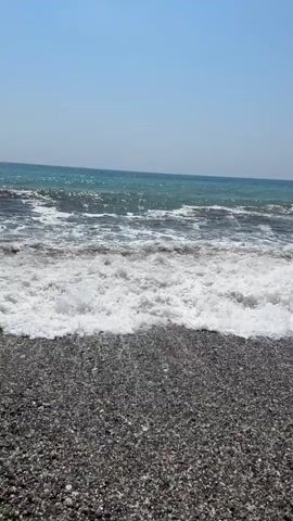 The video shows a close-up view of waves crashing on a dark pebble beach. The water is a deep blue-green, and the waves are breaking with white foam. The sky is clear and blue. The camera is positioned at a low angle, close to the water's edge, giving a sense of immersion in the scene. The footage appears to be taken on a sunny day. There are no people or other objects visible in the frame. The video appears to be a continuous shot with no cuts or edits.