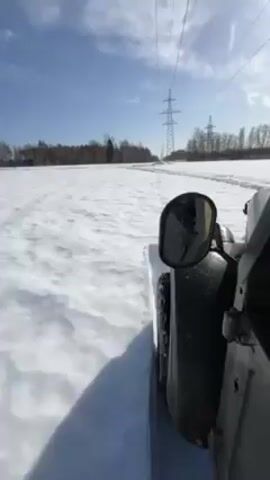 From a low-angle, a vehicle drives across a snow-covered field on a sunny day. The vehicle is dark-colored, and the side mirror is visible on the driver's side.  Power lines run parallel to the vehicle's path. In the distance, a treeline marks the horizon, with a building visible. The sky is blue with sparse clouds. The shadow of the vehicle is visible on the snow.