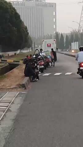 The video shows an outdoor scene with several people and motorcycles parked alongside a road. In the background, there is a large building, construction cranes, and trees. A white vehicle with red markings, possibly an ambulance, is also visible. Some individuals are sitting on a curb next to the motorcycles, while others appear to be standing or walking. The lighting suggests it might be early morning or late afternoon. The environment seems to be a developed area, possibly an industrial or commercial zone, with a road and sidewalk. The overall impression is a casual gathering or break scene.