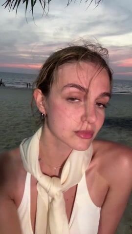 A young woman stands on a beach, blowing a kiss towards the camera, then smiles and poses with her hand under her chin. She has light brown hair pulled back in a messy bun, light skin with some redness on her cheeks, and brown eyes. She is wearing a white halter top and a light beige scarf tied around her neck. A silver ring is visible on her ring finger, and a small tattoo is visible on her forearm. In the background, the beach is sandy with some palm trees and people walking near the water. The sky is a dusky mix of pink, purple, and grey.