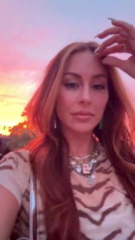 A woman with long red-brown hair, wearing a tan and brown patterned shirt and a layered necklace, is looking directly at the camera and smiling against a backdrop of a vibrant pink and orange sunset. She runs her hand through her hair in the first frame. Palm trees are visible behind her, suggesting a tropical or desert location. People are walking in the background, but they are out of focus. The lighting on her face is a mix of natural sunlight and the warm colors of the sunset.