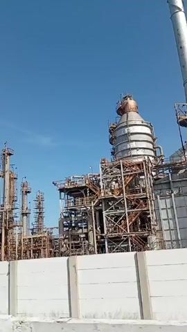 The video shows an industrial complex. The view pans across a series of metal structures, smokestacks, and refinery components against a bright blue sky. The structures appear to be interconnected with scaffolding, pipes, and various industrial equipment. A concrete barrier wall partially obscures the base of the complex. Graffiti or printed messages are visible on the barrier. The scene suggests a large-scale industrial operation, potentially an oil refinery or chemical processing plant.