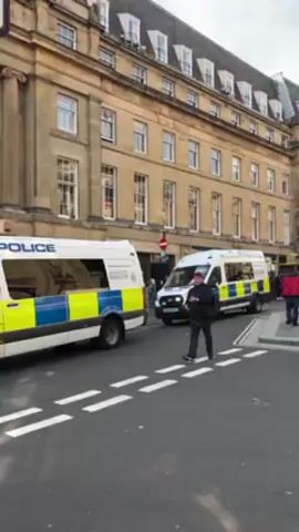 The video shows a street scene in what appears to be a city in the UK, with several police vans parked along the street. The vans are white with blue and yellow markings. A man is standing in the street taking pictures with his phone. Other pedestrians are walking by, including a woman in a red hoodie and two young men. The buildings in the background are several stories tall and appear to be old and well-maintained. There are also signs for taxis and pedestrian walkways. In one part of the video, the camera pans to show more police presence, with a black van parked nearby and numerous people wearing yellow vests walking along the street.