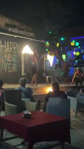 A fire dancer performs on a stage in front of a dark building with "HAPPY NEW YEAR" written on it. The dancer is shirtless and wears dark shorts, holding a flaming staff in each hand, spinning them in a circular motion. Another man in dark clothes stands behind the fire dancer, near a small controlled fire on the stage. To the right of the stage, a third man is crouching. Several people are seated at tables in the foreground, watching the performance. The area is illuminated by the fire and various colorful lights.