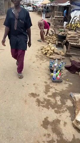 A woman in a pink shirt and printed shorts is sorting through a pile of yams on the side of a street. She's barefoot and moving the yams around, potentially arranging them. Other vendors' stalls line the street, with items like bananas and pineapples visible. Bottled drinks are also on display nearby. The road appears to be unpaved and has some puddles. Other people are walking and riding motorcycles on the street. The setting appears to be a market or a street-side market in a populated area.