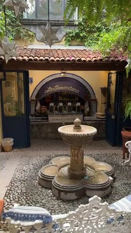 A medium shot shows a courtyard featuring "La Samaritana Barra De Aguas Frescas". A short, ornate stone fountain with multiple bowl-shaped tiers sits in the center, surrounded by decorative rocks. The bar is set against a wall with open doors on either side. Above the bar is an arched sign bearing the business name. The roof has brown tiles and there are star-shaped decorations hanging from above. The courtyard is framed by greenery. No people are visible.