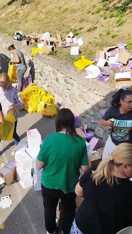 A group of people are gathered outdoors, unloading items from the back of a silver van. The scene appears to be in a rural setting, possibly a village street, with houses and a grassy hillside visible in the background. The van's rear hatch is open, and people are either taking items out or placing them down.

Several individuals are holding bright yellow plastic bags, suggesting they are carrying supplies. There are also numerous cardboard boxes and some purple boxes scattered around on the asphalt and a low stone wall. Some of the boxes are open, revealing white tissue paper or packaging. In the foreground, a brown cardboard box contains bananas.

The activity suggests a distribution or donation event, possibly of food or goods, given the presence of the yellow bags and food items. The people are dressed casually, indicating a daytime event. The lighting suggests it is daytime, with sunshine casting shadows. The overall atmosphere seems to be one of cooperative effort. The location is identified as Oeștii Ungureni, Romania.